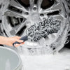A hand is using a super plush wheel cleaning brush to clean a car wheel covered in soap foam.