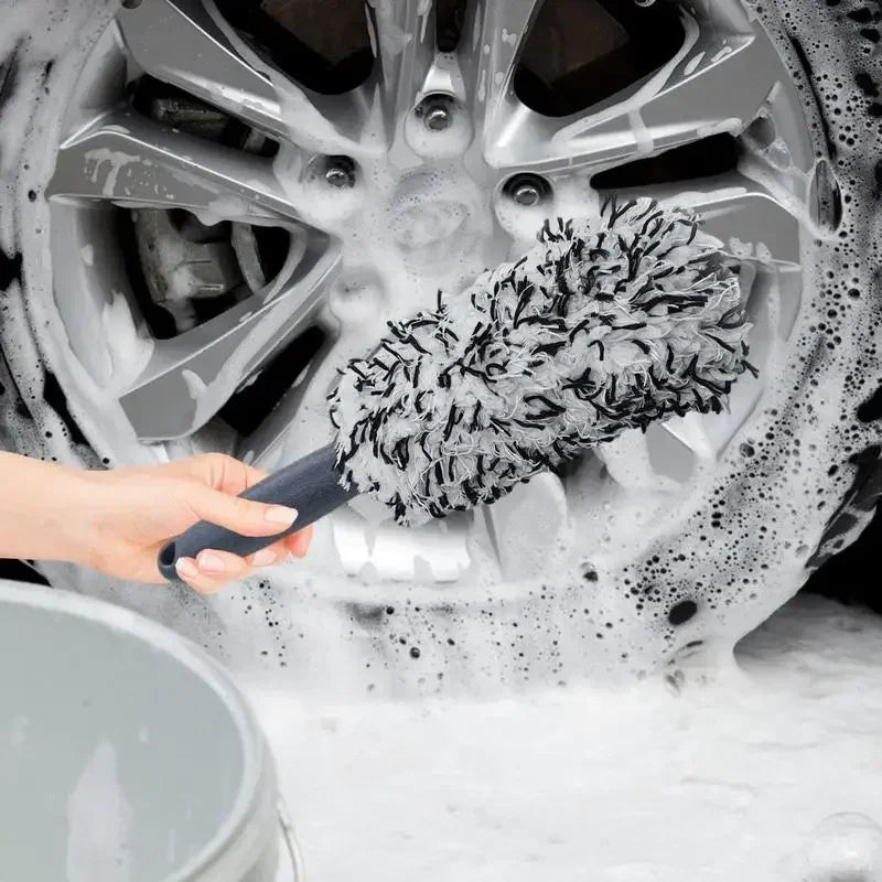 A hand is using a super plush wheel cleaning brush to clean a car wheel covered in soap foam.