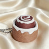 A fidget keychain shaped like a rolled cake with brown frosting and white swirl details resting on satin fabric.