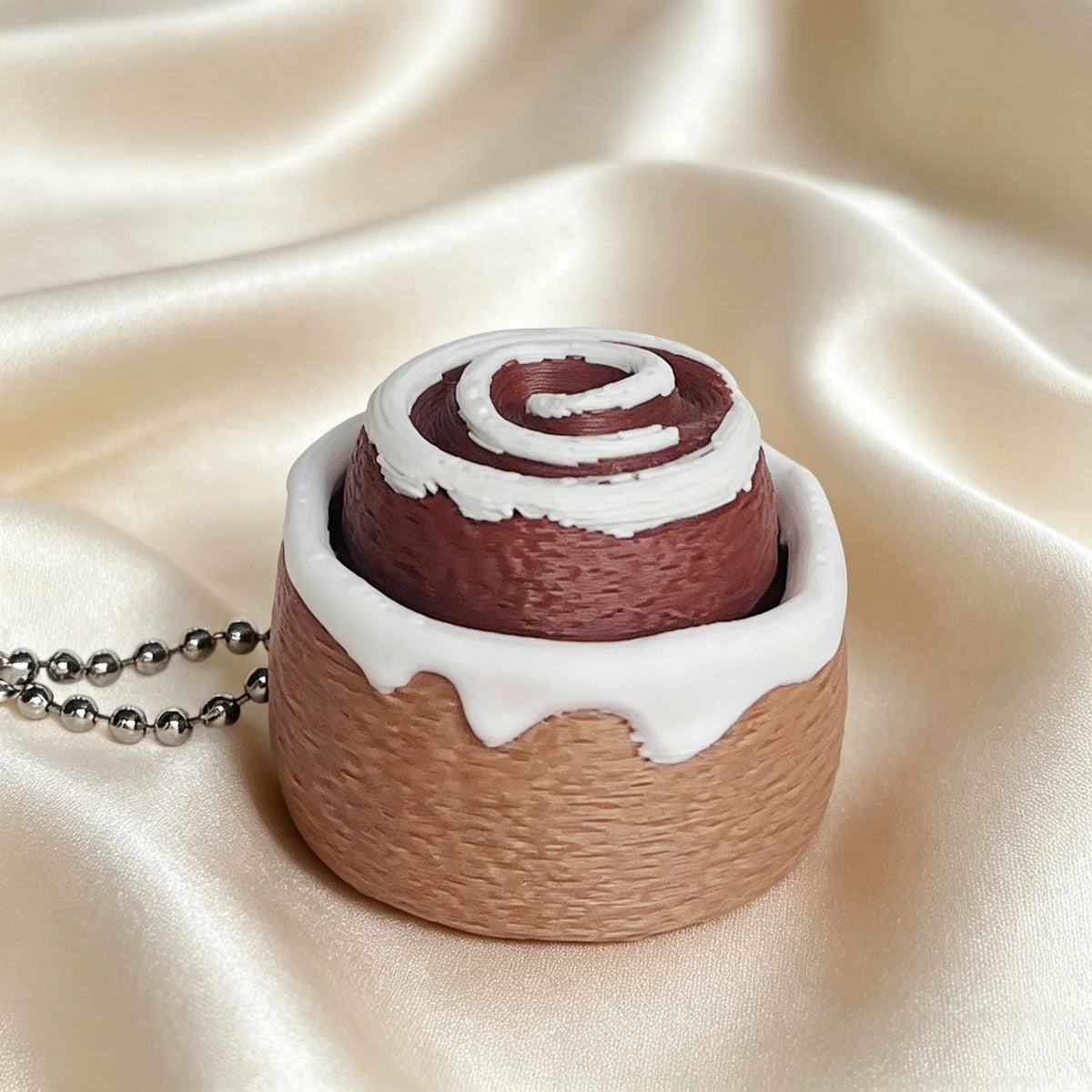 A fidget keychain shaped like a rolled cake with brown frosting and white swirl details resting on satin fabric.