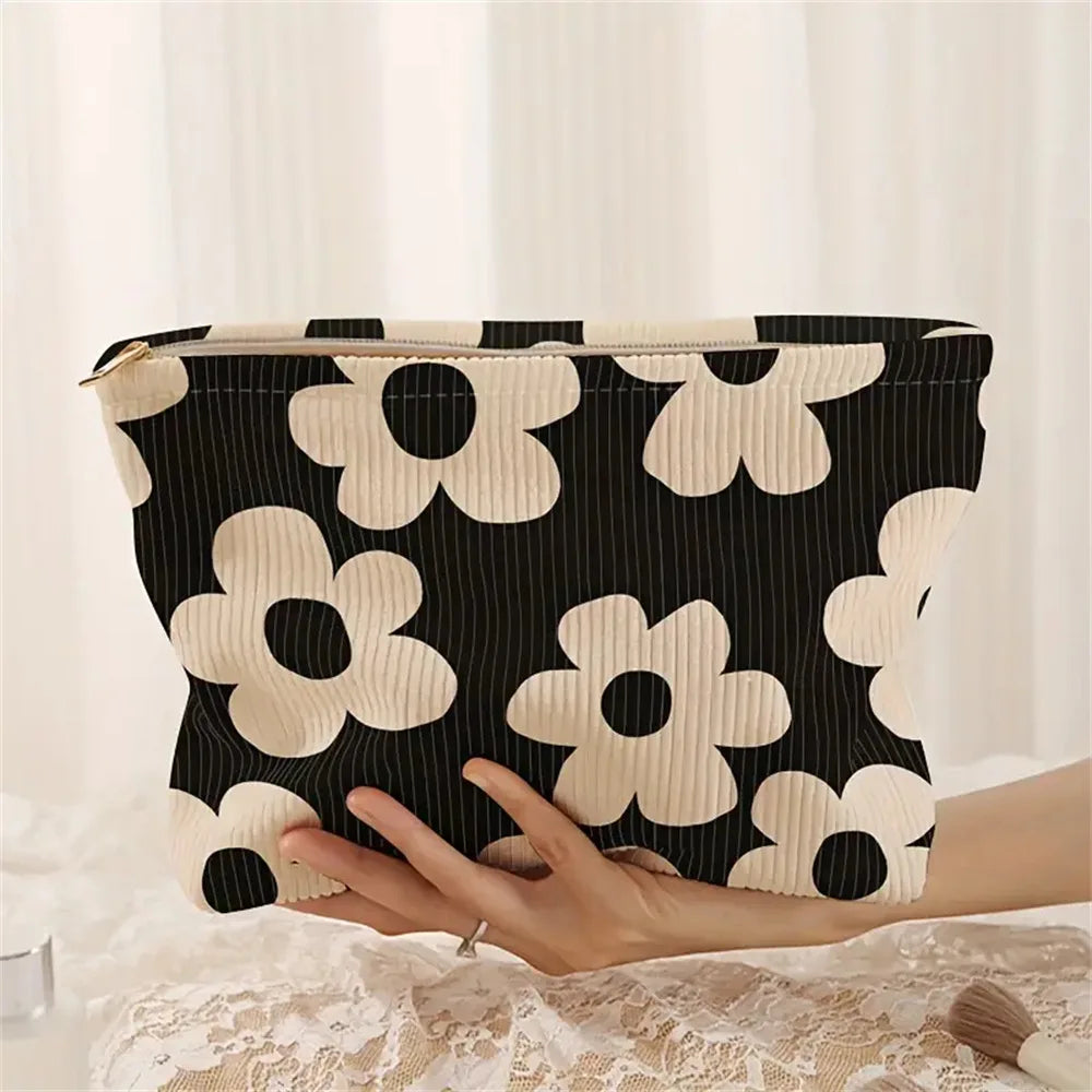 A beige floral corduroy makeup bag with a black and white pattern is held by a hand on a white lace surface.