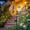 A solar owl garden light with moon decor hangs over a staircase surrounded by flowering plants.