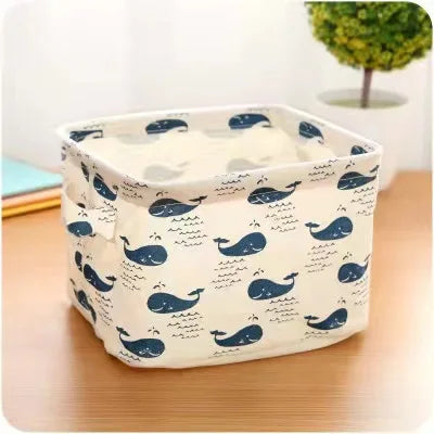 A white cotton and linen storage basket with handles featuring a pattern of blue whale silhouettes on a wooden surface.