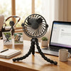 A black flexible octopus USB fan mounted on a tripod stand sits on a wooden desk next to a laptop and a mug.