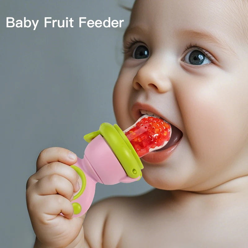 A baby is using a pink and green rotating silicone fruit feeder to feed a piece of fruit.