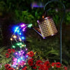 A vintage solar watering can garden light with a shepherd's hook illuminates a vine of colorful LED lights over red flowers.
