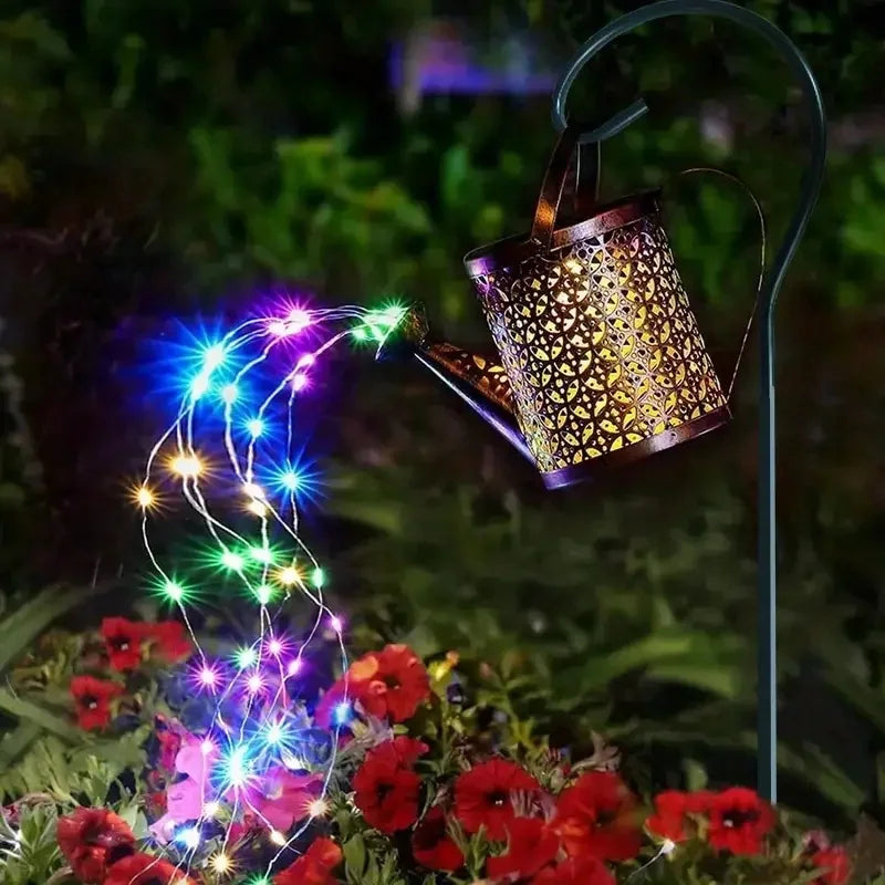 A vintage solar watering can garden light with a shepherd's hook illuminates a vine of colorful LED lights over red flowers.