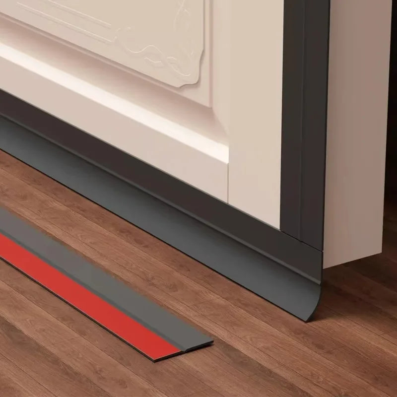 A door draft stopper with a red and gray strip is shown installed under a door frame on a wooden floor.