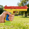 A hand holds a red and yellow building block toy pistol with shell ejecting action in an outdoor setting.