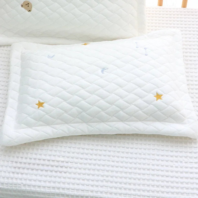 A white quilted baby pillow with gold stars and blue crescent moons resting on a white textured fabric.
