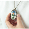 A hand holds a silver floral embroidery necklace featuring blue and orange flowers on a white background.
