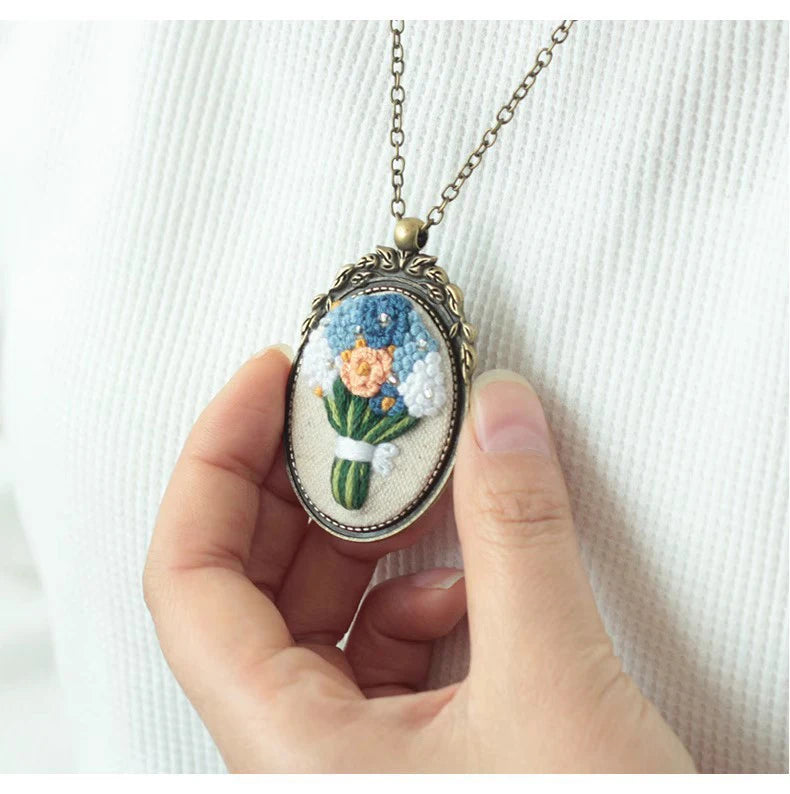 A hand holds a silver floral embroidery necklace featuring blue and orange flowers on a white background.