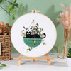 A circular embroidery hoop displaying a black cat and floral designs on white fabric, set against a green background.