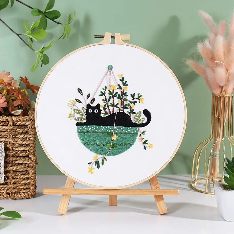 A circular embroidery hoop displaying a black cat and floral designs on white fabric, set against a green background.