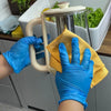 A hand wearing blue nitrile gloves holds a yellow sponge over a clear glass pitcher in a white sink.