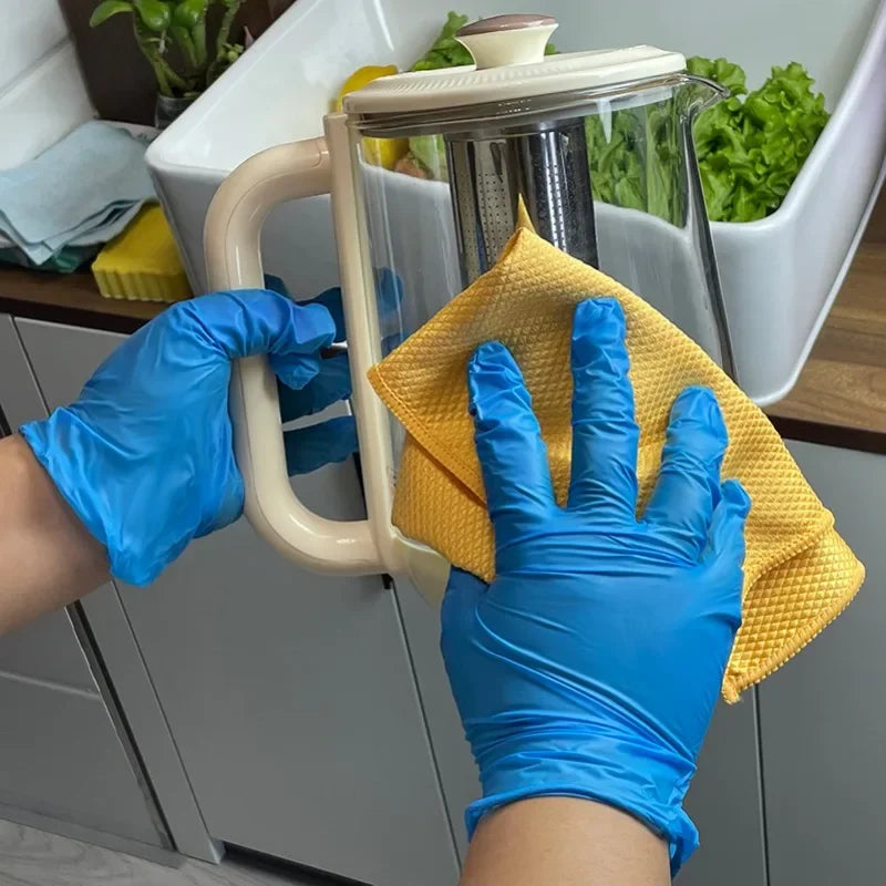 A hand wearing blue nitrile gloves holds a yellow sponge over a clear glass pitcher in a white sink.
