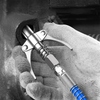 A hand holds a heavy-duty grease gun coupler with a silver metal body and blue tip.