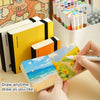 Hands open a yellow sketchbook filled with watercolor paintings and an array of art markers.