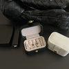 A black leather jewelry travel case is shown open, displaying several rings and a separate light beige storage box.