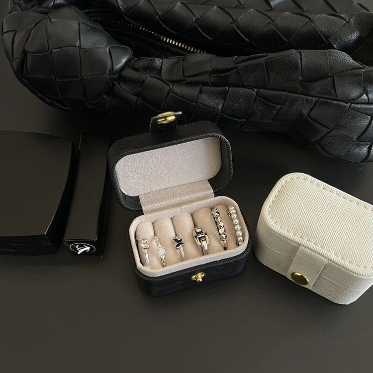 A black leather jewelry travel case is shown open, displaying several rings and a separate light beige storage box.