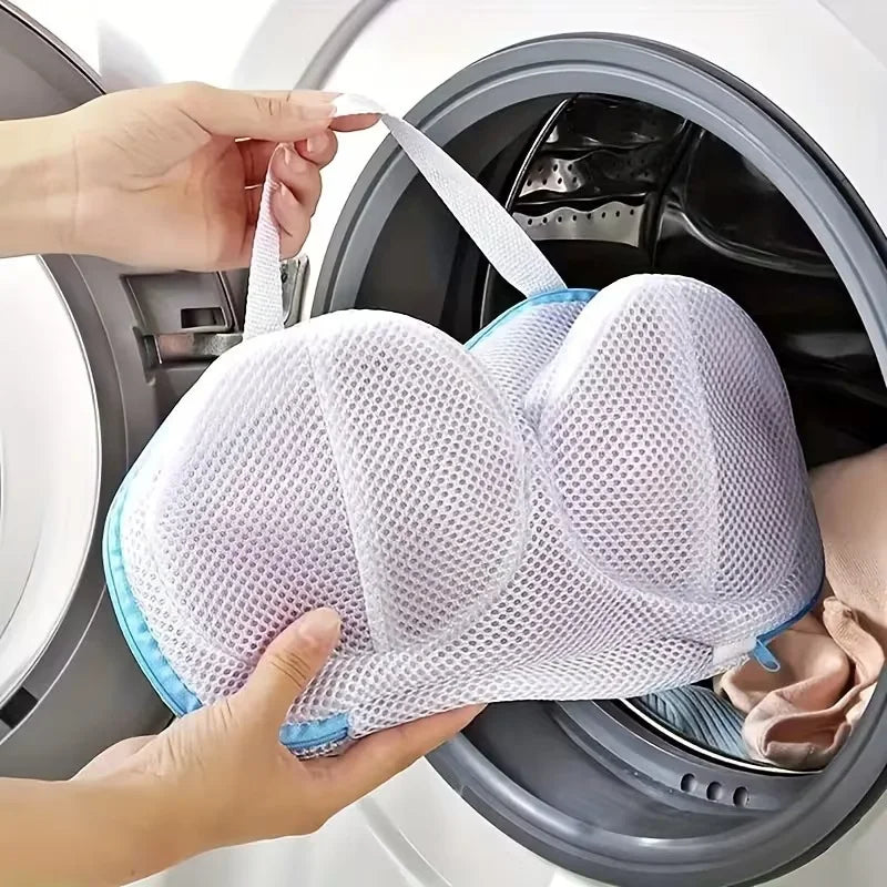 A hand is placing a white mesh laundry bag for bras and lingerie into the drum of a washing machine.