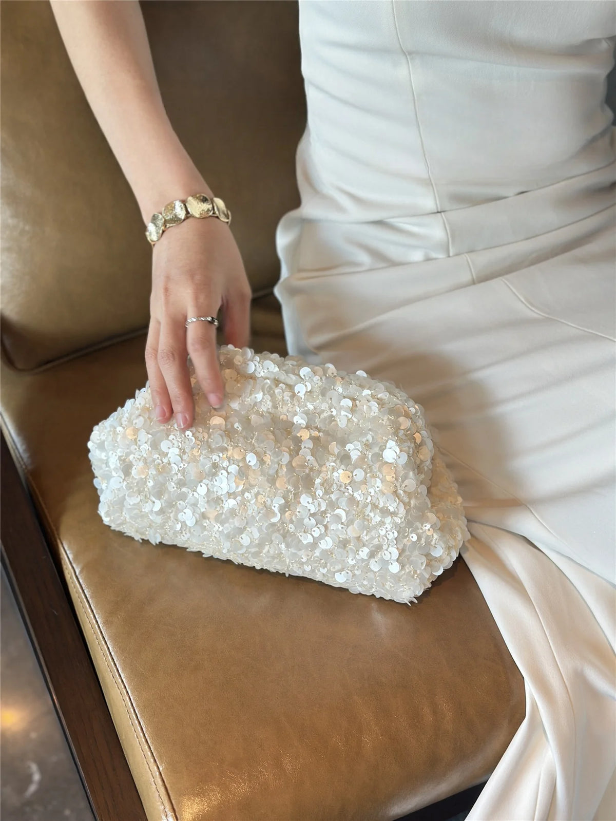 A hand places a white sequin beaded chain shoulder clutch bag on a brown leather sofa.