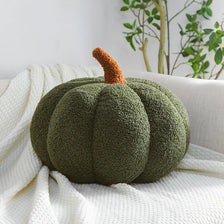A round, moss-green plush pumpkin pillow with an orange stem resting on a white knitted fabric.