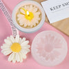A silicone mold shaped like a daisy flower is shown next to a candle and a pink silicone mold.