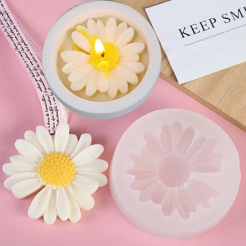 A silicone mold shaped like a daisy flower is shown next to a candle and a pink silicone mold.