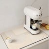 A white electric tea maker sits on a beige counter mat with striped patterns, set against a light background.