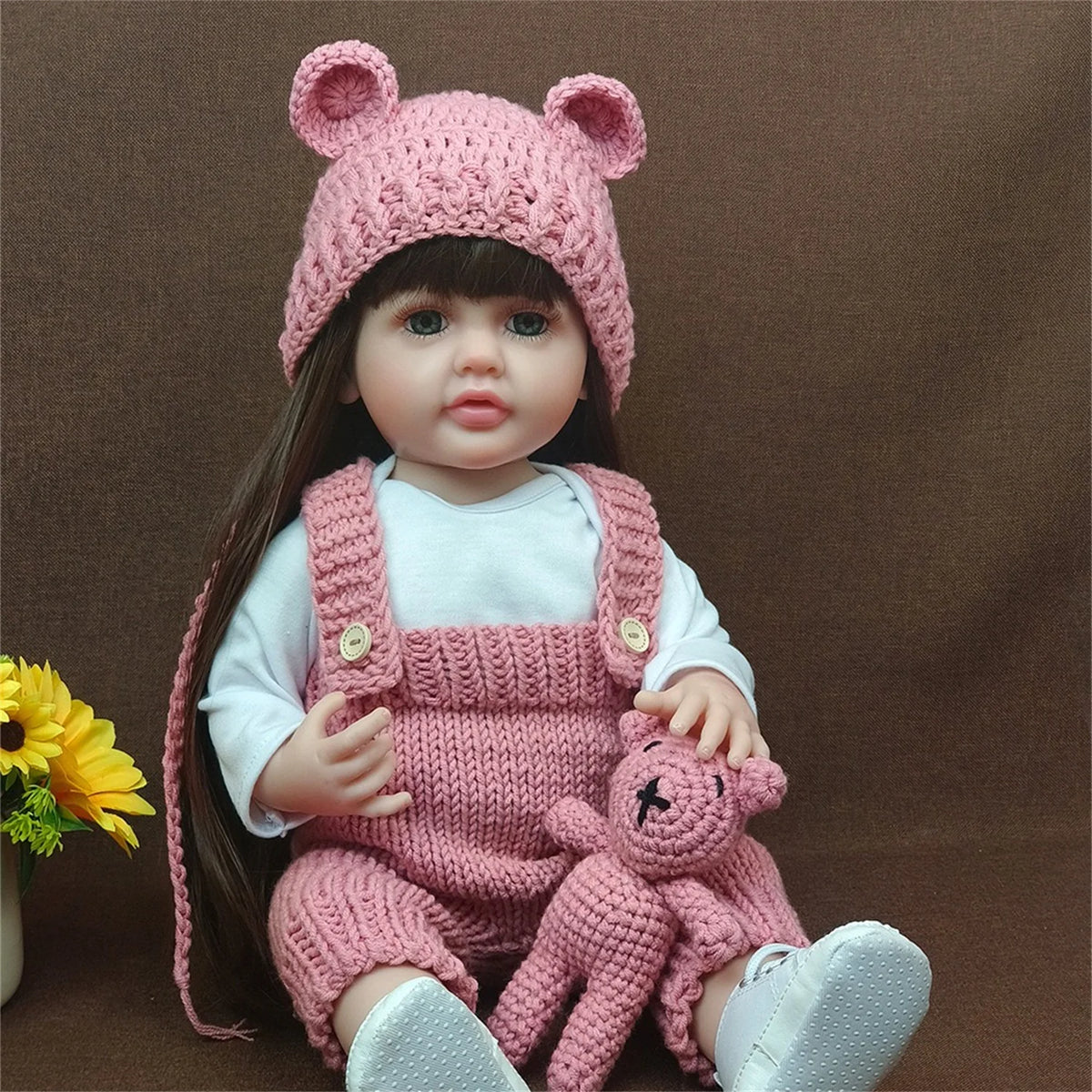 A pink silicone baby doll wearing a matching knitted hat and overalls sitting on a brown background.