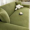 A textured olive green stretch jacquard sofa cover with a cream decorative pillow and a cup on the seat.