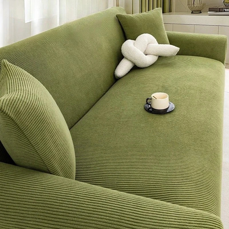 A textured olive green stretch jacquard sofa cover with a cream decorative pillow and a cup on the seat.