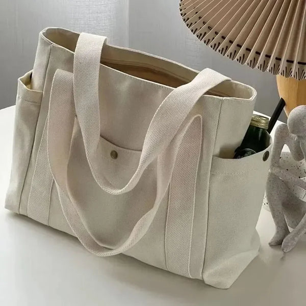 Una bolsa de tela beige con tiras de color beige claro junto a una botella oscura y un sombrero tejido.