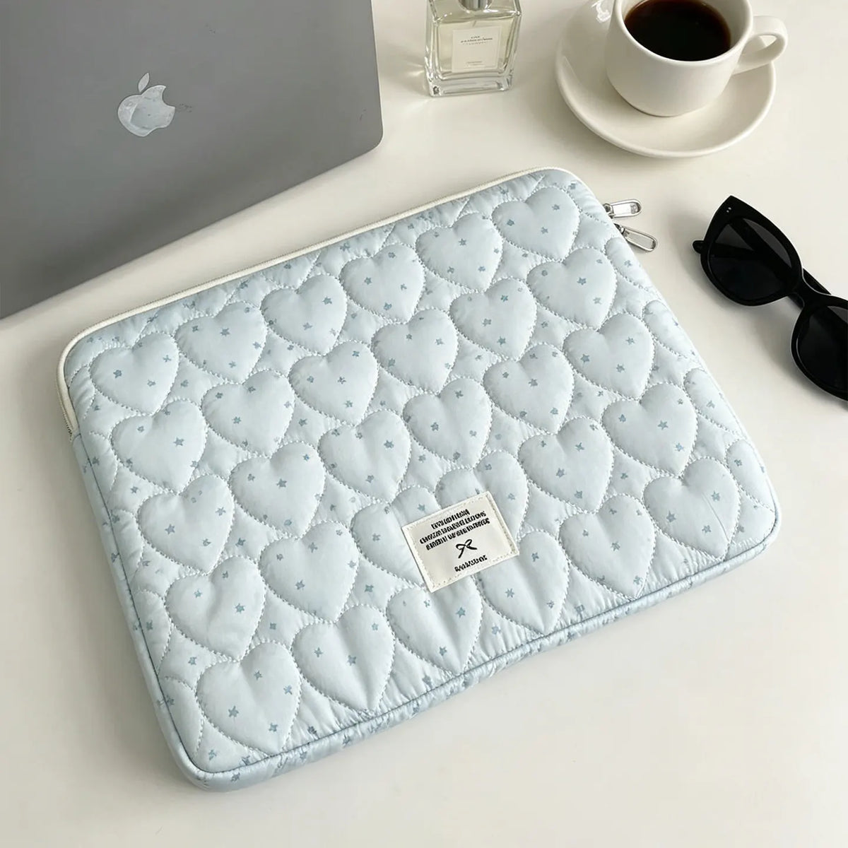 A light blue quilted laptop sleeve with a diamond pattern resting on a white surface next to a silver perfume bottle and sung