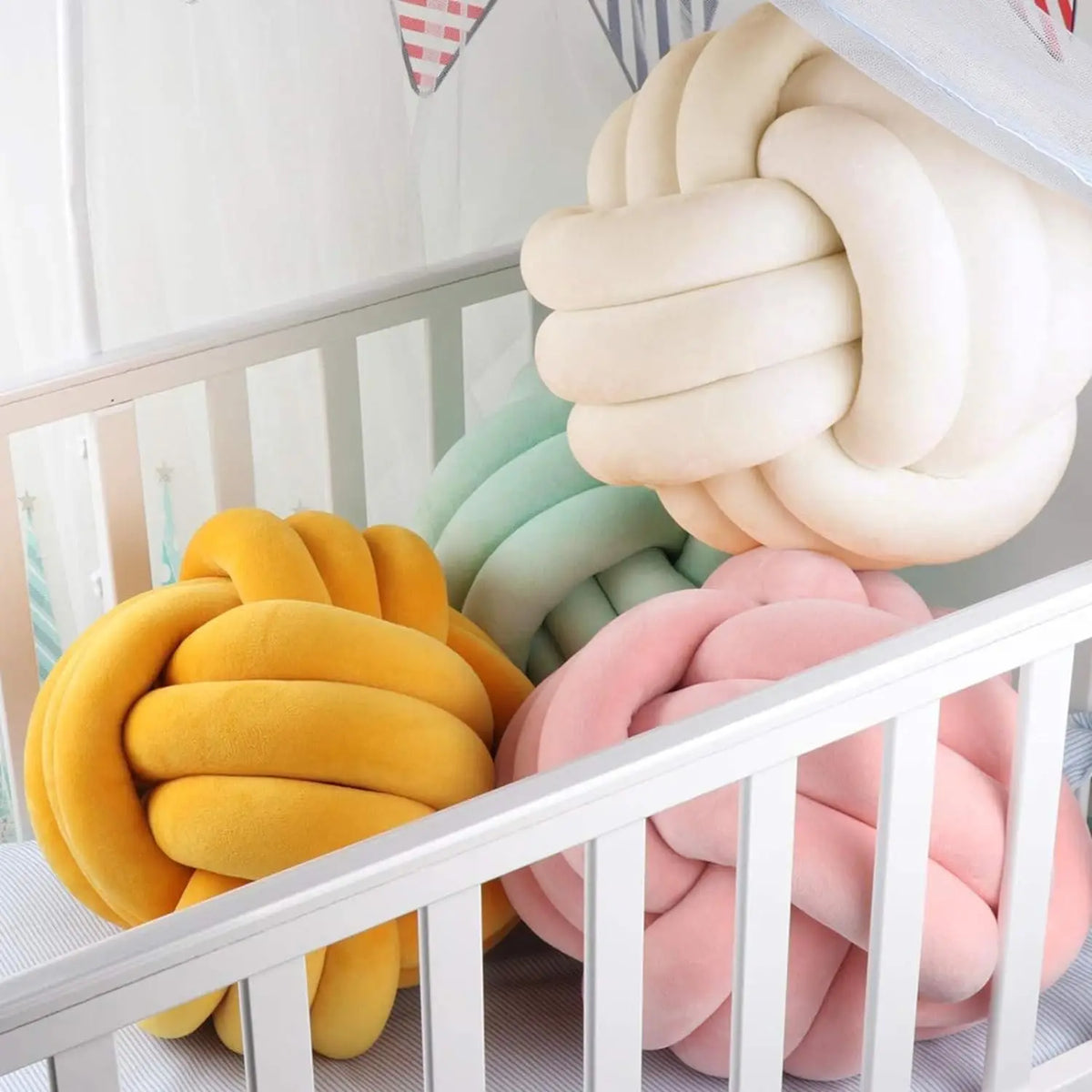 A collection of soft, round knitted velvet throw pillows in yellow, mint green, and pink resting on a white crib railing.