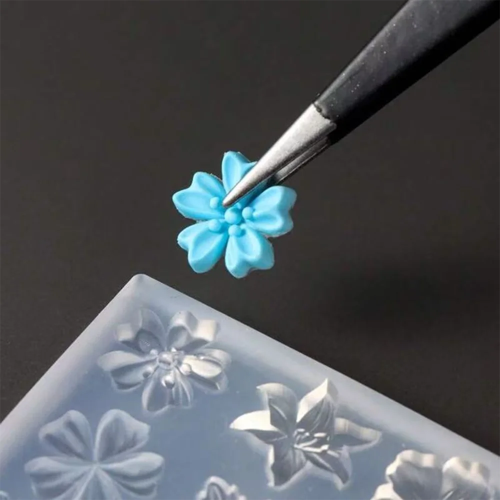 A fine black tool is applying a small, light blue silicone flower detail onto a clear nail art mold.
