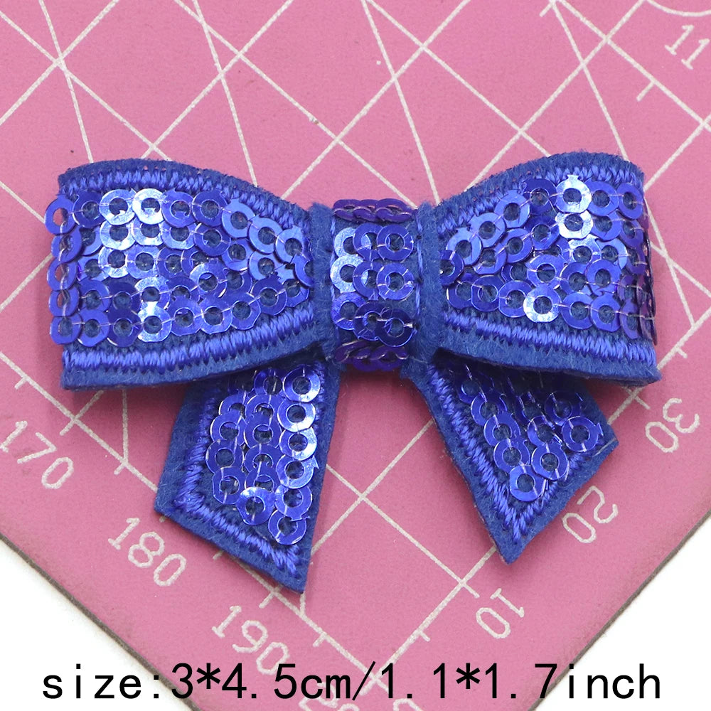A royal blue sequin bow patch for DIY crafts displayed on a pink grid background.