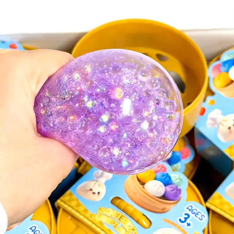 Eine Hand hält ein lila glitzerndes Squishy-Dumpling-Sensorspielzeug in einem gelben Behälter.