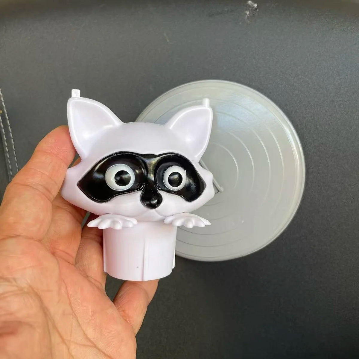 A white cat-shaped toilet paper holder with black markings is held against a clear plastic lid.