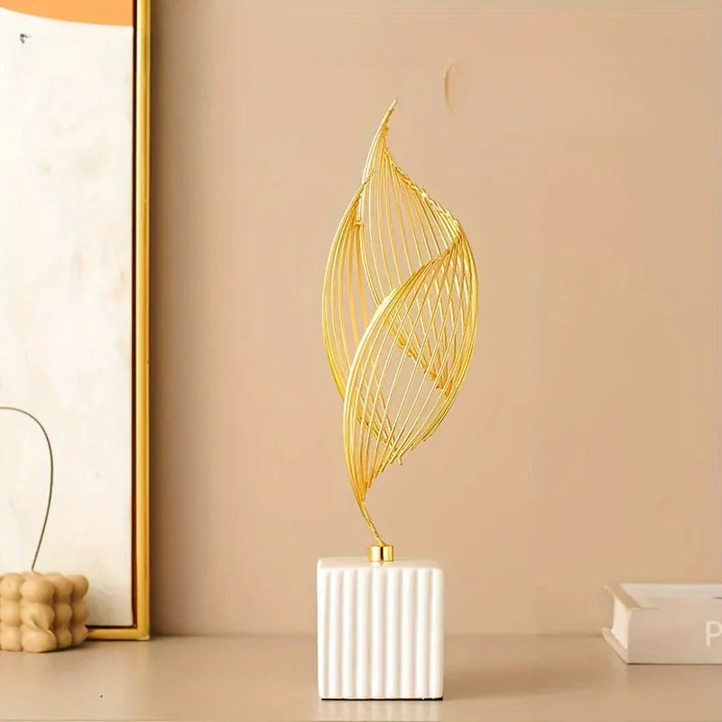 A golden, leaf-shaped sculpture stands on a white ceramic block against a neutral background.
