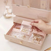 A hand is opening a light pink jewelry storage box containing multiple clear self-sealing bags for rings, necklaces, and brac
