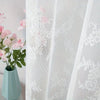 White lace curtain panel with floral embroidery and sheer white fabric next to a pink flower arrangement in a light blue vase