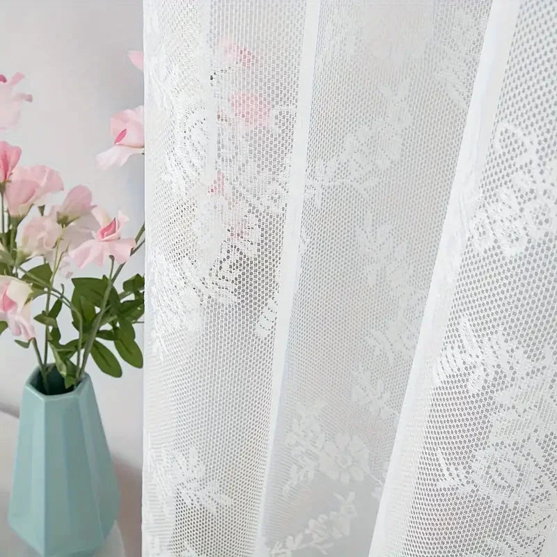 White lace curtain panel with floral embroidery and sheer white fabric next to a pink flower arrangement in a light blue vase