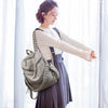 A woman wearing a beige top and dark skirt holds an elegant soft leather backpack with silver rivets.