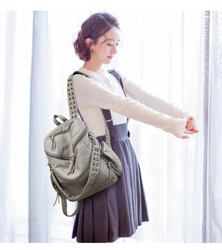 A woman wearing a beige top and dark skirt holds an elegant soft leather backpack with silver rivets.