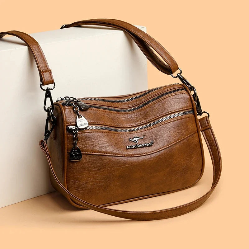 A brown leather compact square crossbody bag with a gold-tone chain and leather strap resting on a light background.
