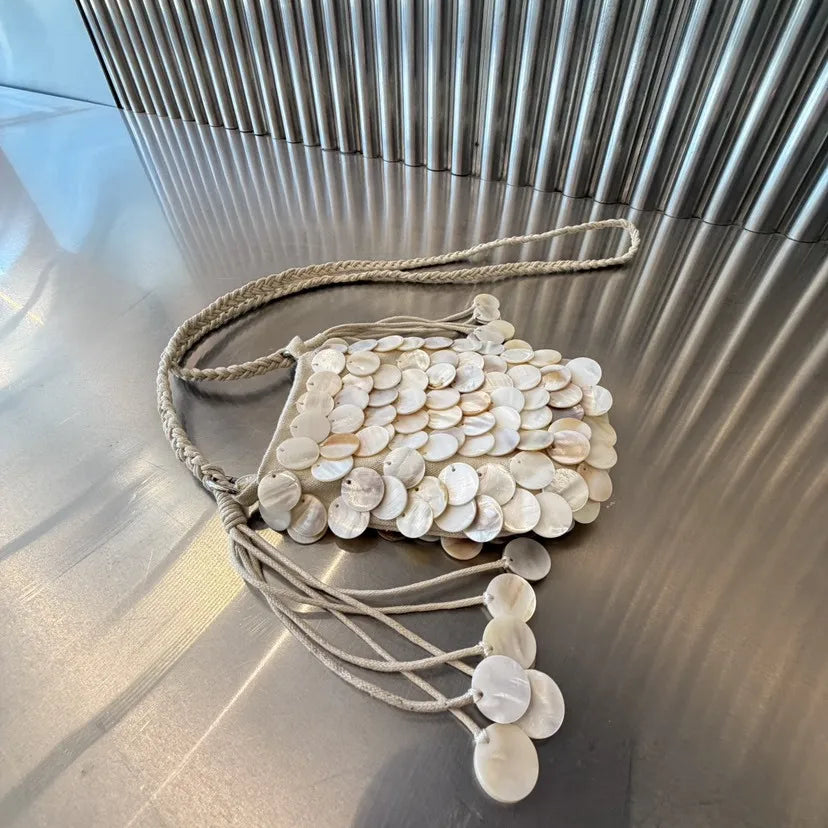 A beige sequin accent chain shoulder bag with a woven strap resting on a silver metallic surface.