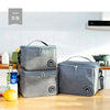 Three grey insulated lunch bags with shoulder straps are displayed on a wooden table in a bright kitchen setting.