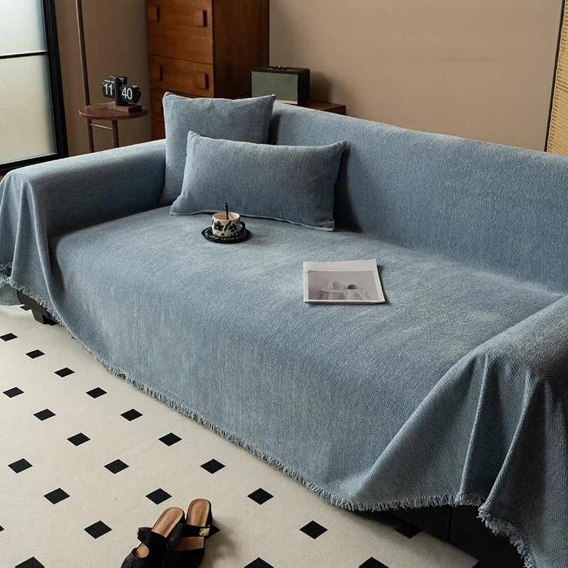 A light blue herringbone chenille sofa throw draped over a sofa with matching pillows and a decorative tassel edge.